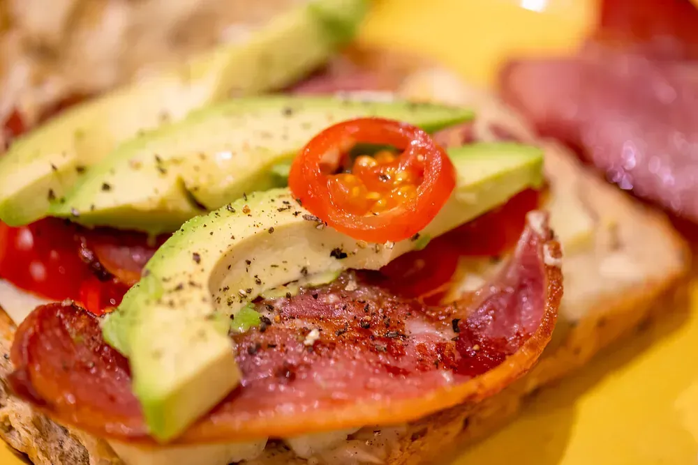 Un primer plano de un sándwich con aguacate, tocino, tomates y pimientos en un plato amarillo.