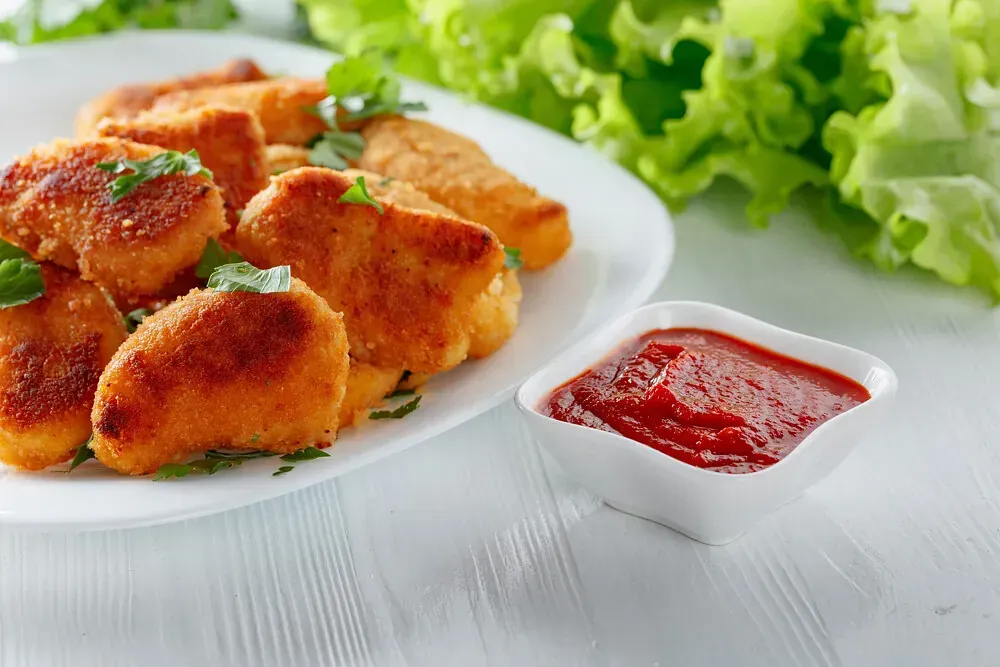 Un plato blanco cubierto con nuggets de pollo frito junto a un tazón de salsa de tomate.