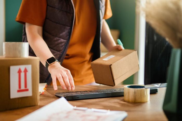 Una persona empaqueta una caja de cartón en una mesa de envío con cinta adhesiva y una computadora portátil.