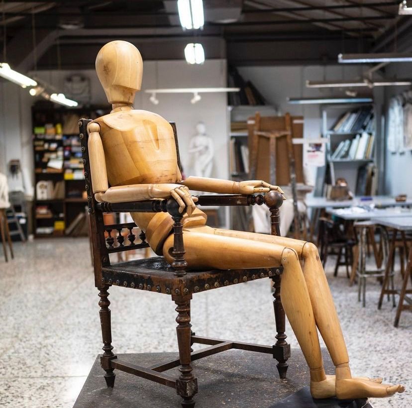 Maniquí de artista de madera sentado en una silla oscura y ornamentada en un estudio, rodeado de materiales de arte y muebles.