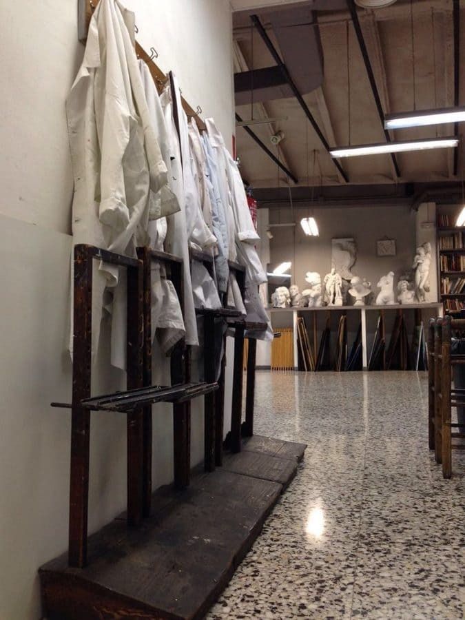 Batas blancas de arte cuelgan de un perchero en un estudio de arte con estatuas y suelo de baldosas.