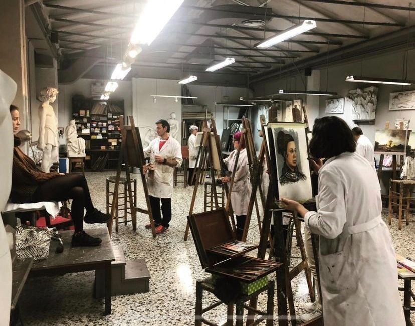 Clase de arte: estudiantes con batas blancas pintan en caballetes, algunos con retratos. La modelo posa sobre una plataforma.