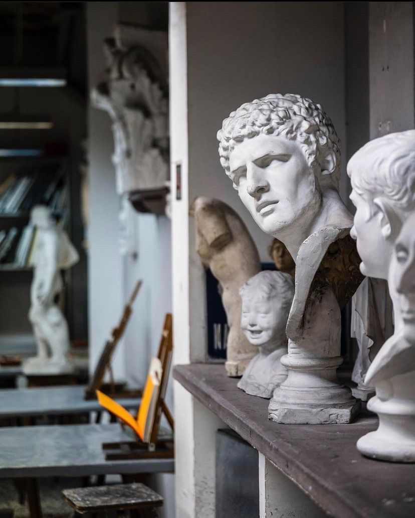 Estudio de escultura: bustos de yeso sobre un estante. Al fondo, una figura y un espacio de trabajo con taburetes y un caballete.