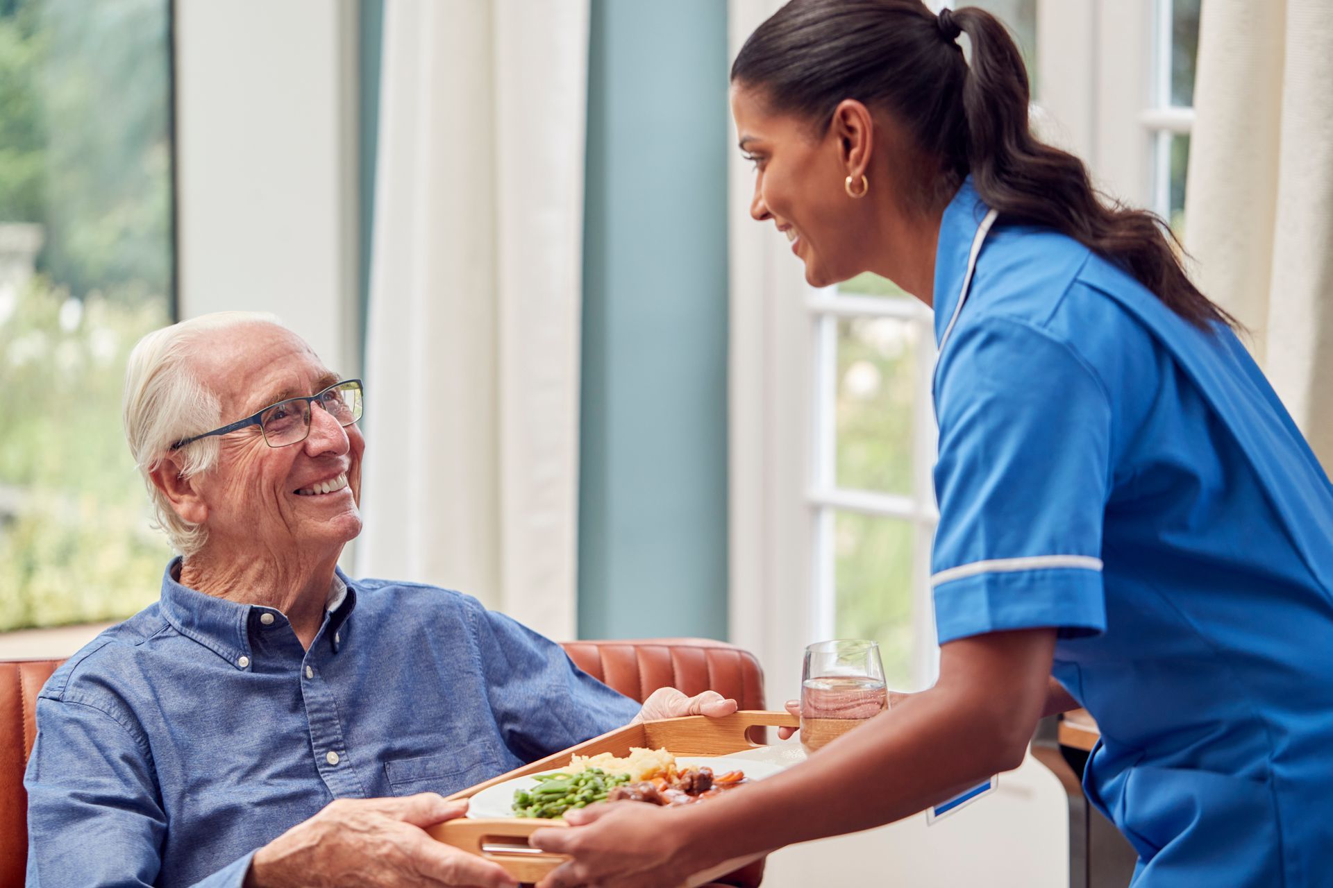 Une infirmière en uniforme bleu sert un repas à un patient souriant, assis dans un fauteuil près d'une fenêtre.