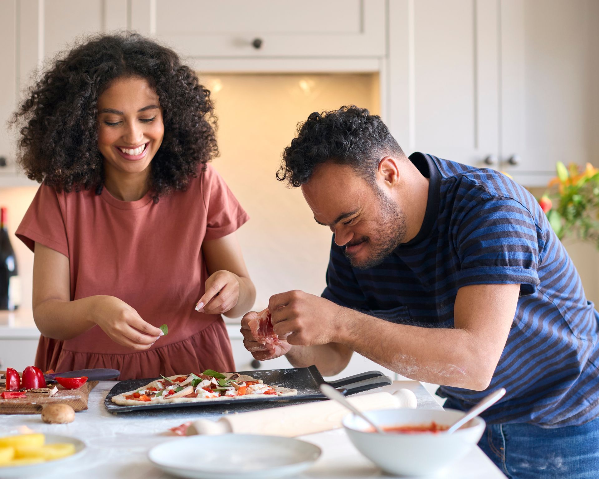 Deux personnes sourient en préparant des pizzas ensemble dans une cuisine lumineuse.
