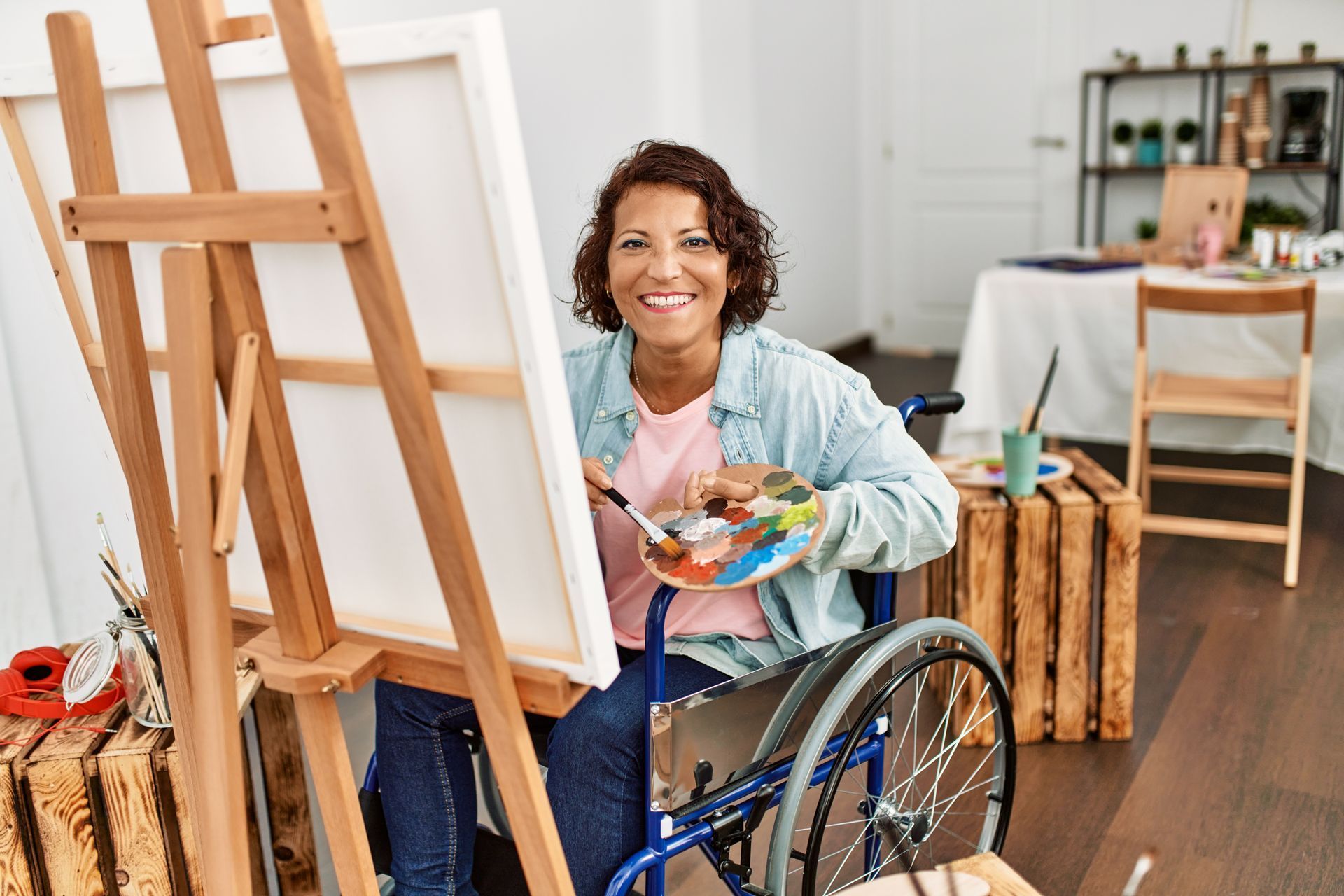 Une femme en fauteuil roulant peint sur un chevalet dans un atelier lumineux, souriante et tenant une palette.