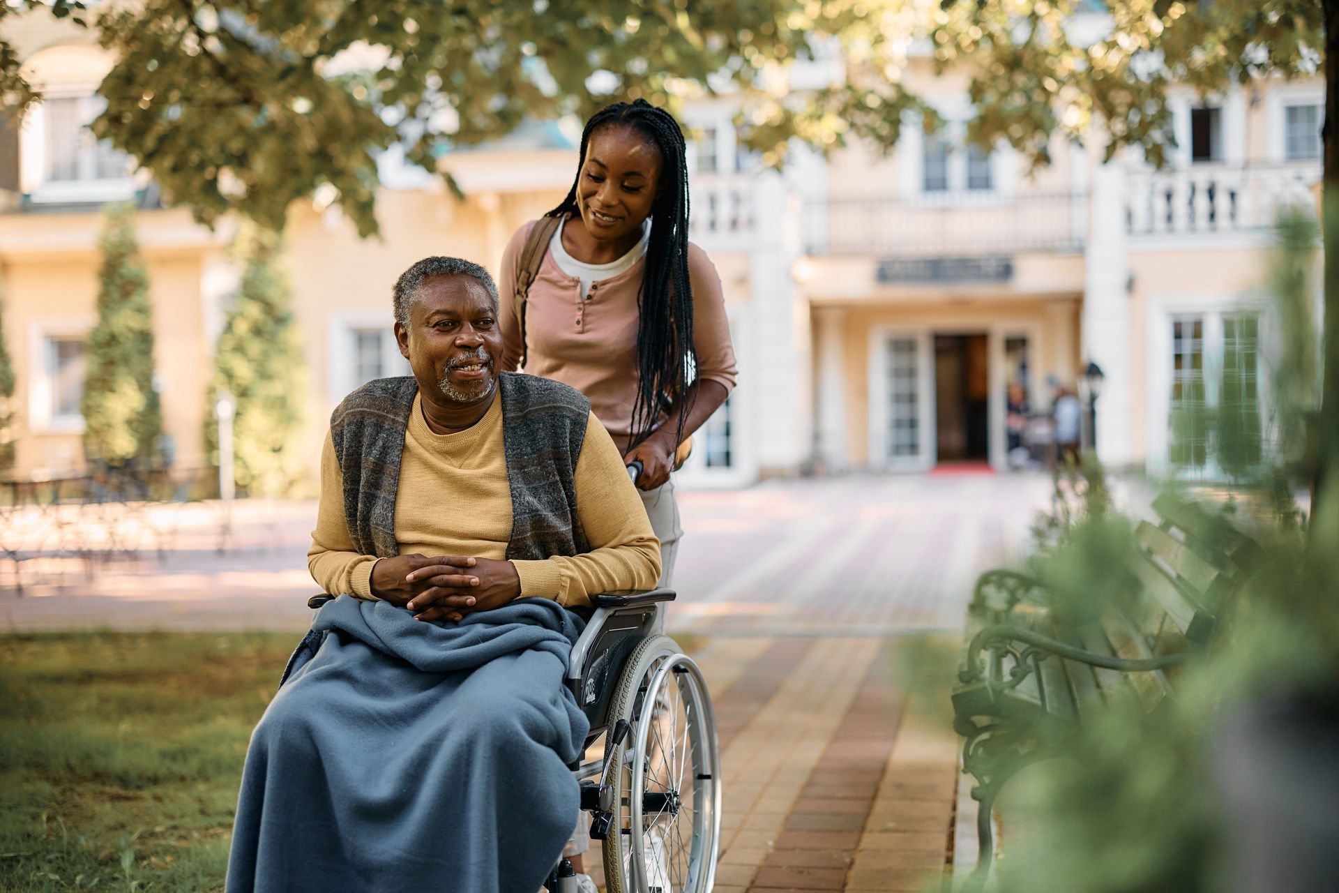 Un homme souriant dans un fauteuil roulant à l'extérieur, une femme penchée sur lui, sur une allée du campus