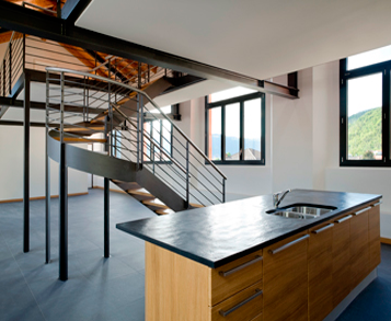 Interior de loft moderno con escalera de caracol, isla de cocina y grandes ventanales con vista al exterior.