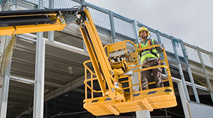 Trabajador de la construcción en plataforma elevadora amarilla, con casco y chaleco de seguridad, trabajando en el exterior del edificio.