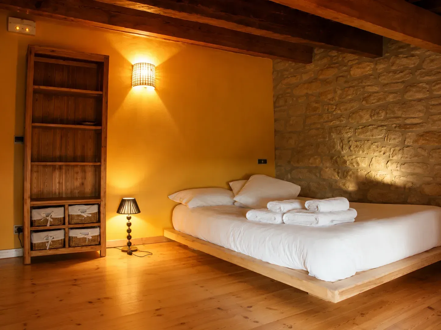 Acogedor dormitorio rústico con suelo de madera, cama blanca, pared de piedra e iluminación cálida con lámparas.