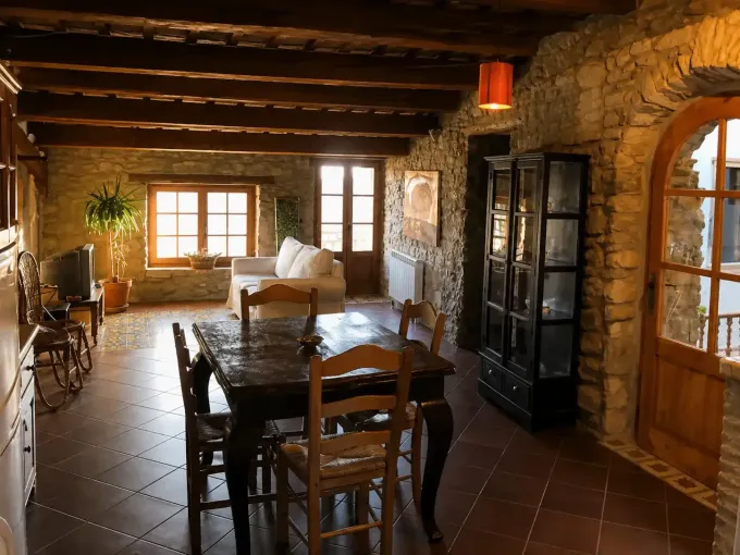 Salón rústico de piedra con vigas de madera, suelo de baldosas, mesa de comedor, sillas y grandes ventanales.