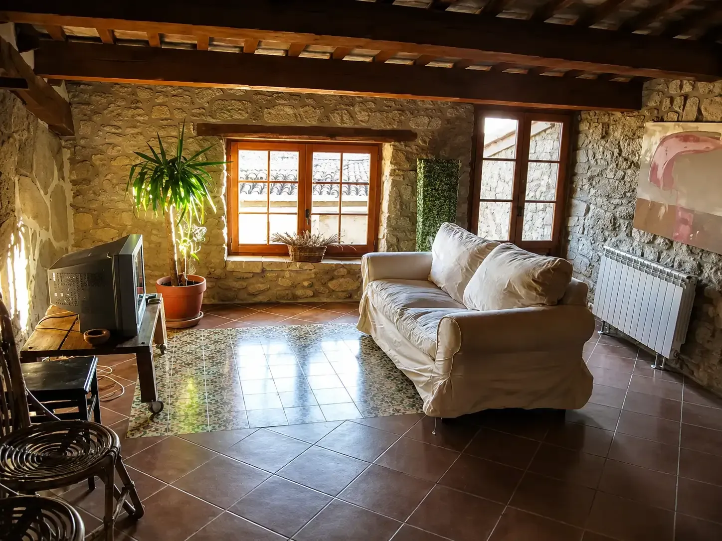 Sala de estar rústica de piedra con suelo de baldosas, sofá, ventana y planta en maceta, bañada por la cálida luz del sol.