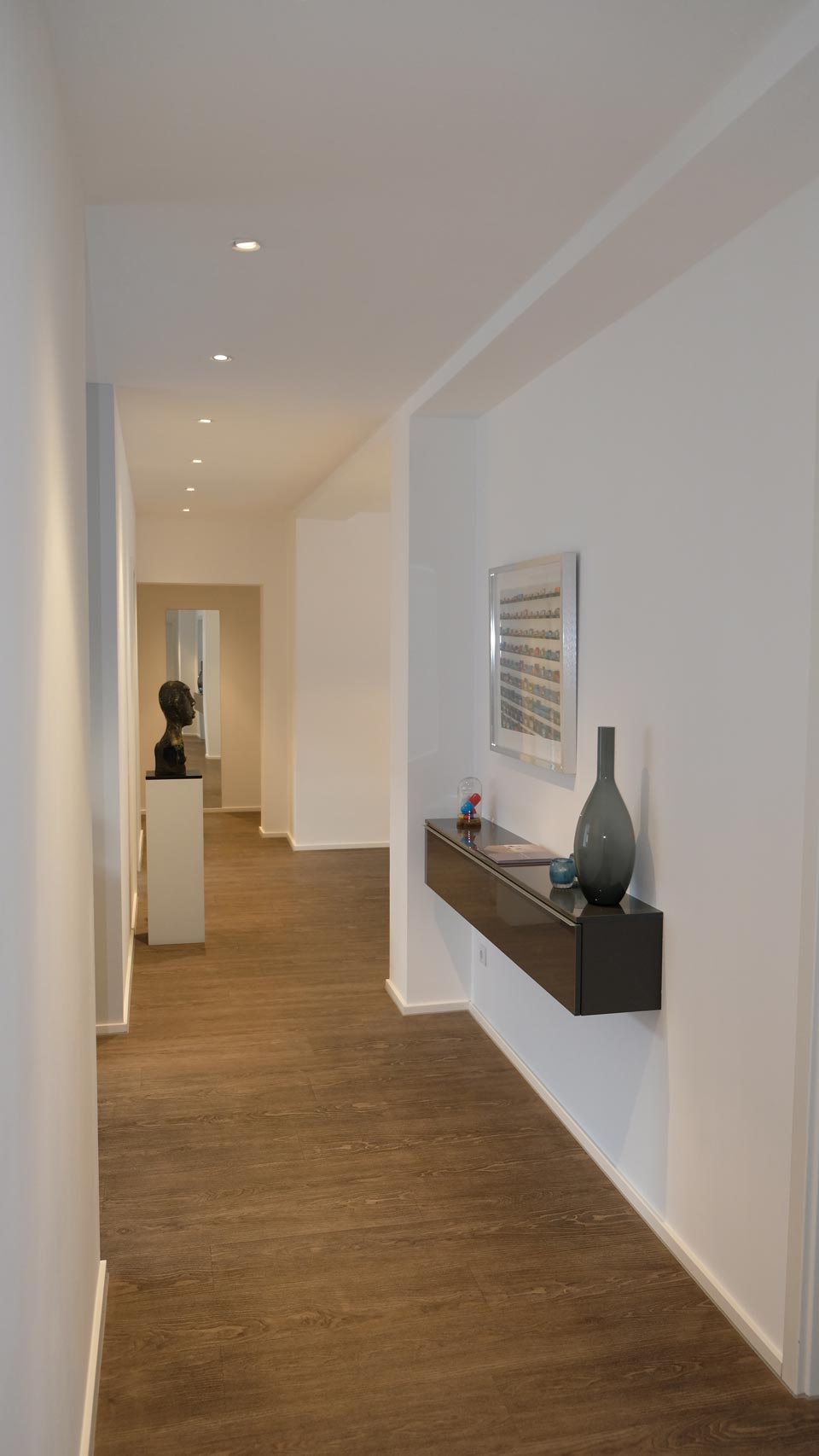 Langer Flur mit weißen Wänden, braunem Boden und minimalistischem Dekor, darunter eine Skulptur und ein schwebendes Regal mit einer Vase.