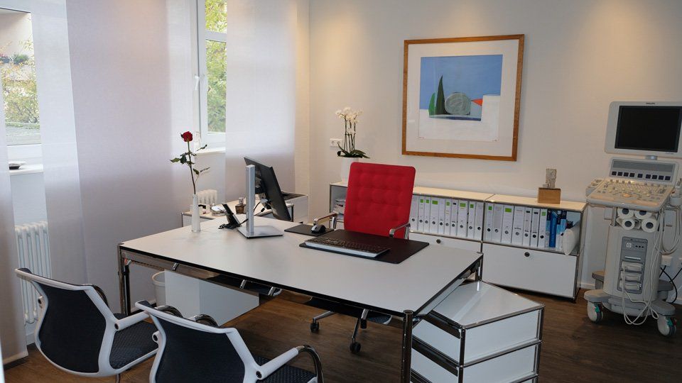Arztpraxis mit Schreibtisch, Stühlen, Ultraschallgerät und Kunstwerken. Weiße und rote Akzente.