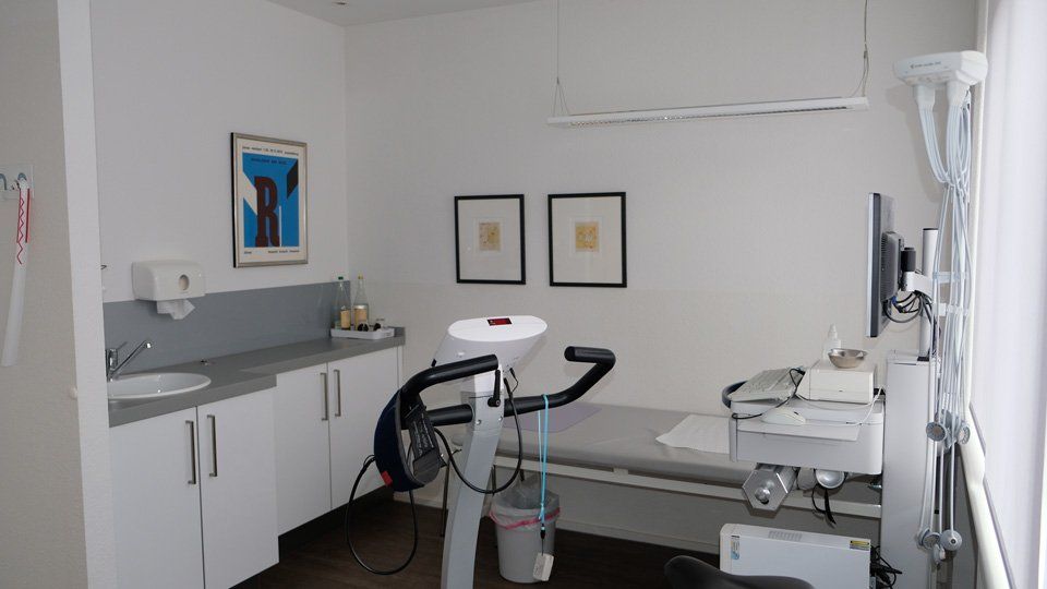 Medizinischer Untersuchungsraum mit Heimtrainer, Waschbecken und Untersuchungsliege.