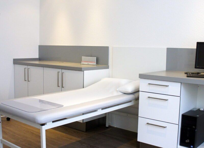 Untersuchungsraum mit Krankenbett, Schränken und Computer. Farbschema in Weiß und Grau.