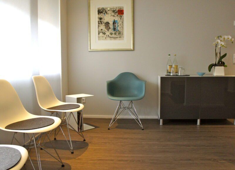 Wartezimmer mit Stühlen, Schrank, Kunstwerken und Blumen.