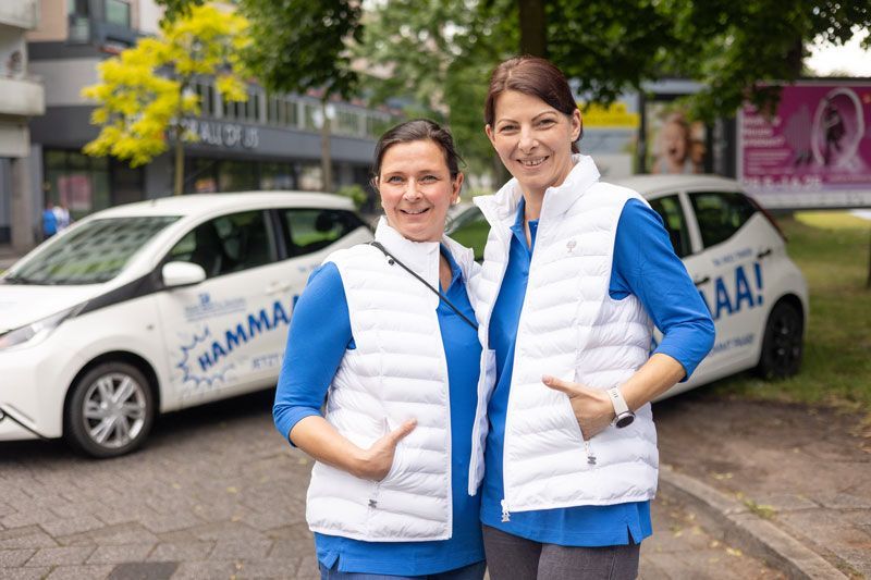 Zwei Frauen in blauen Oberteilen und weißen Westen lächeln neben einem weißen Markenauto.