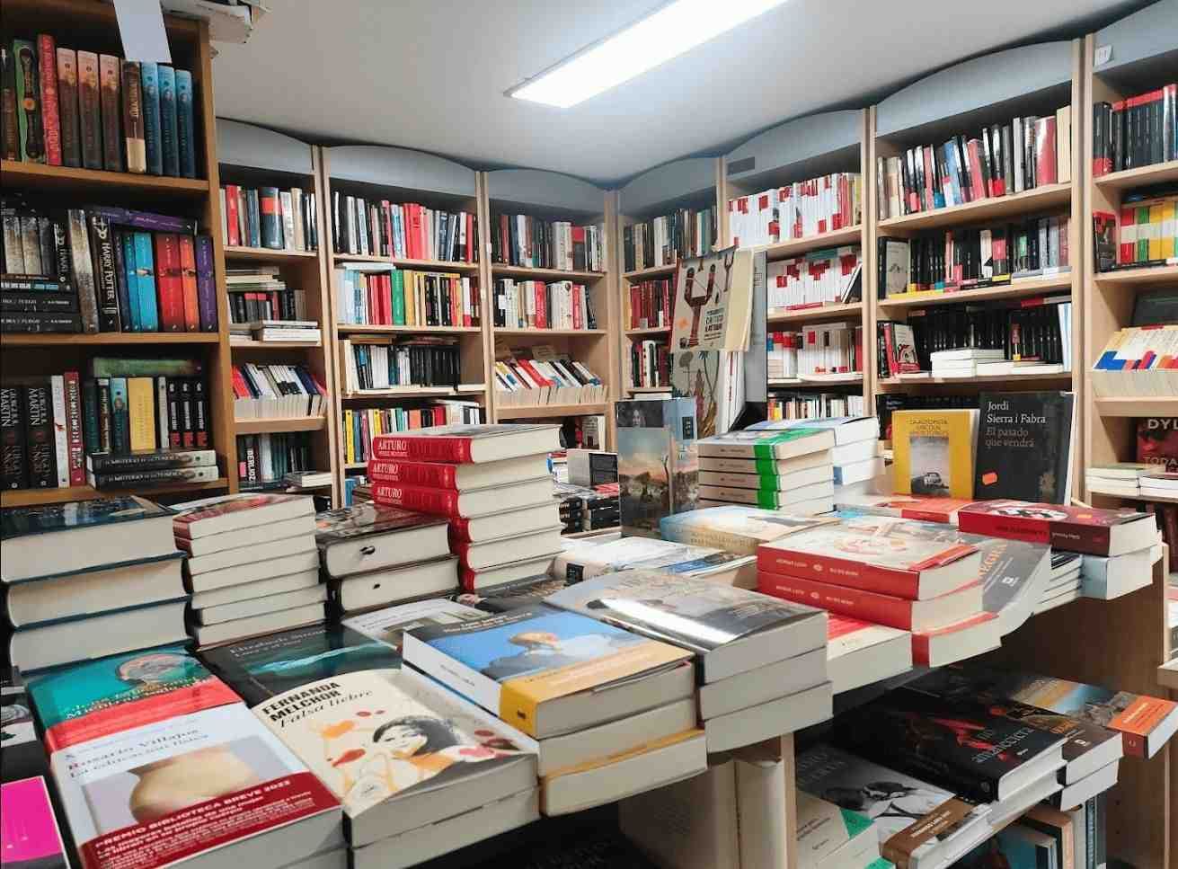 Pequeña librería abarrotada de pilas de libros sobre mesas y estantes, bajo brillantes luces fluorescentes.