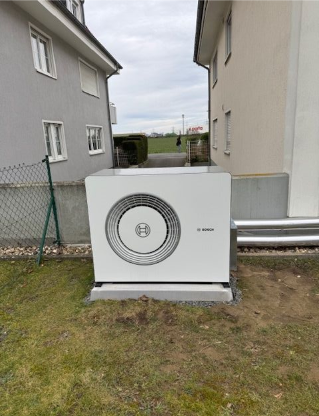 Wärmepumpe neben zwei Häusern mit freiliegenden Rohren und einem umzäunten Garten