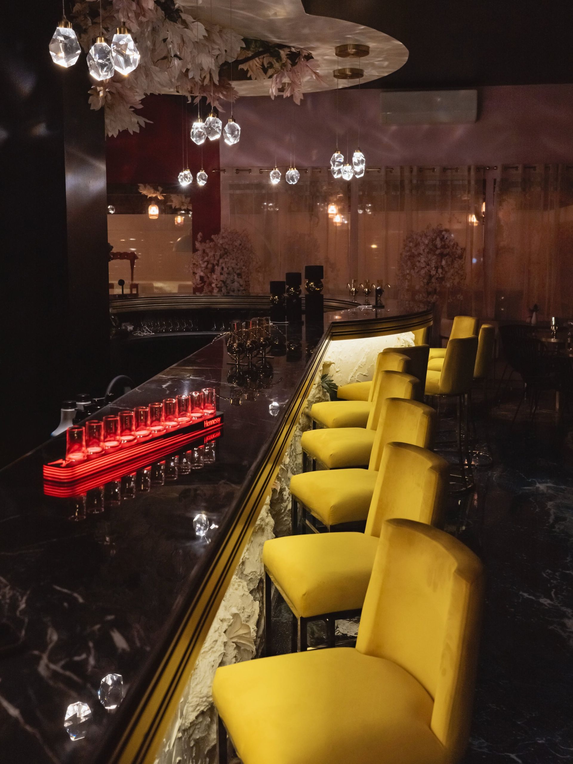 Tabourets de bar jaunes autour d'un élégant comptoir sombre, éclairé par des lumières rouges indirectes et des suspensions lumineuses en verre.