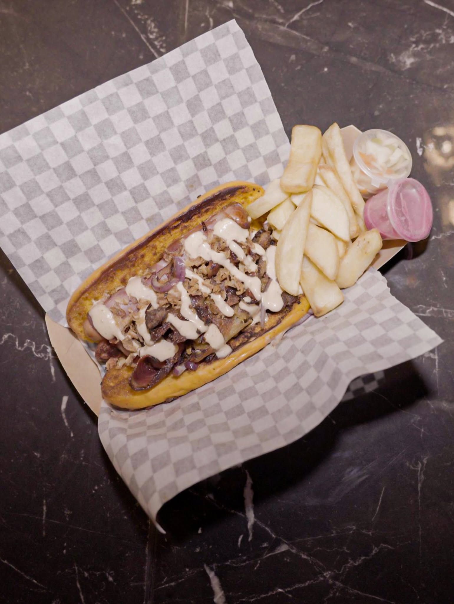Sandwich grillé avec sauce, frites et cornichons tranchés, servi sur du papier à carreaux sur une table sombre.