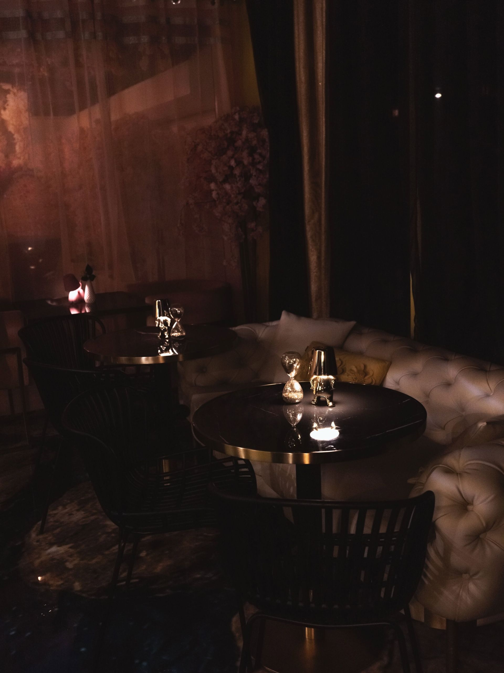 Salon tamisé avec tables rondes, fauteuils capitonnés et bougies près d'une fenêtre le soir.