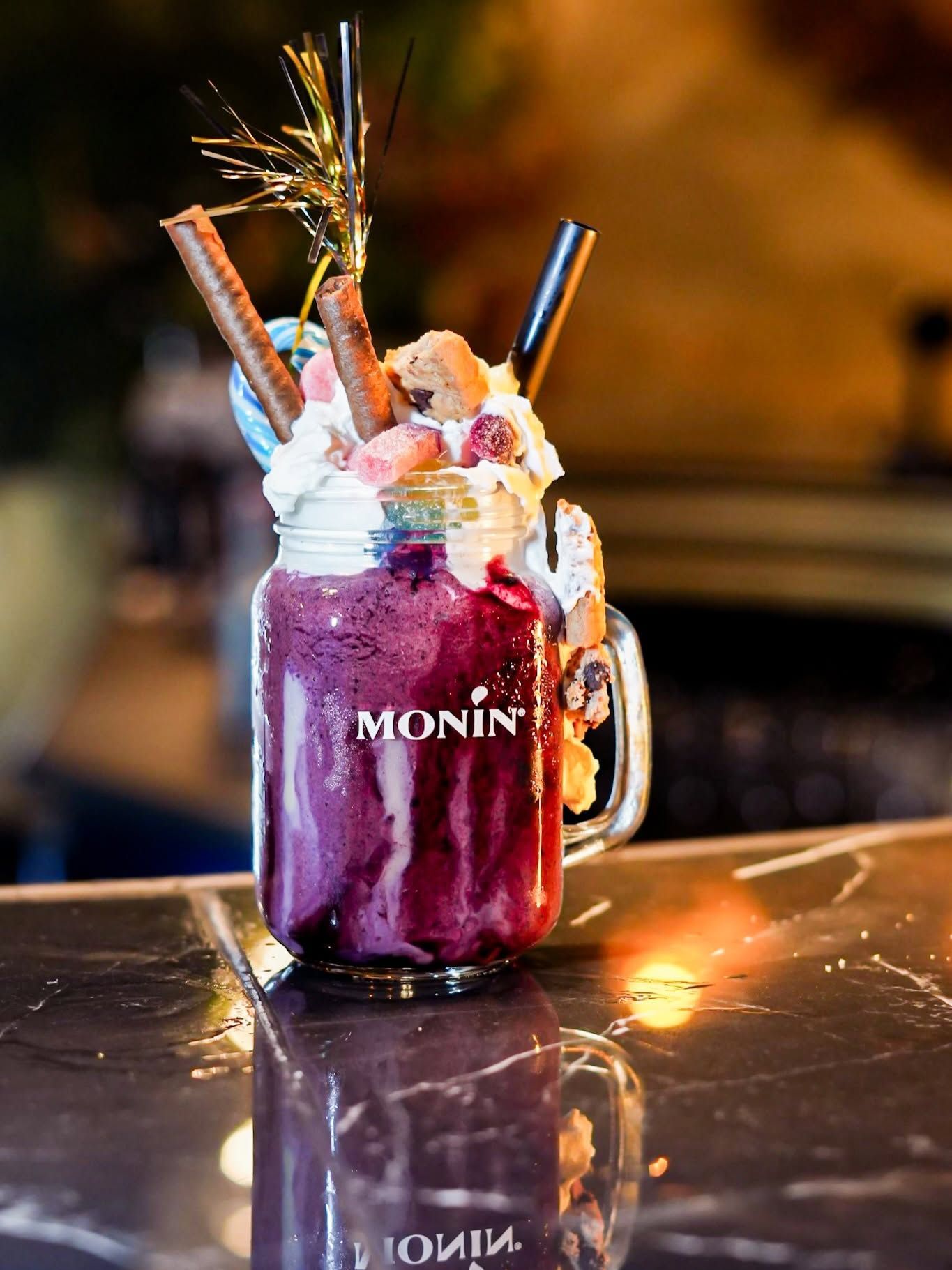 Cocktail violet servi dans un bocal Monin avec de la crème fouettée, des fruits et des pailles, sur un comptoir de bar