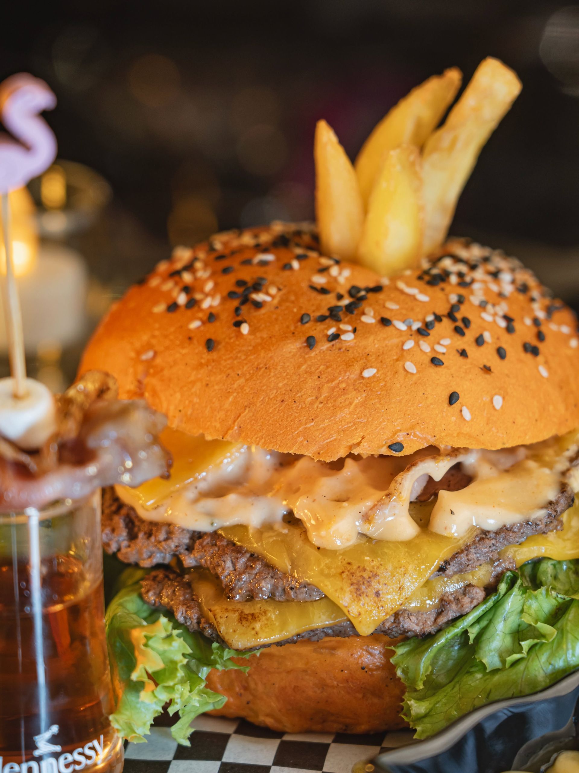 Gros plan sur un hamburger au sésame garni de frites, de fromage fondu, de laitue et accompagné d'une boisson.