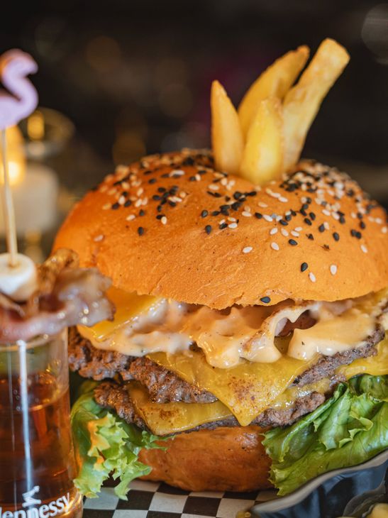 Gros plan sur un hamburger au sésame garni de frites, de fromage fondu, de laitue et accompagné d'une boisson.