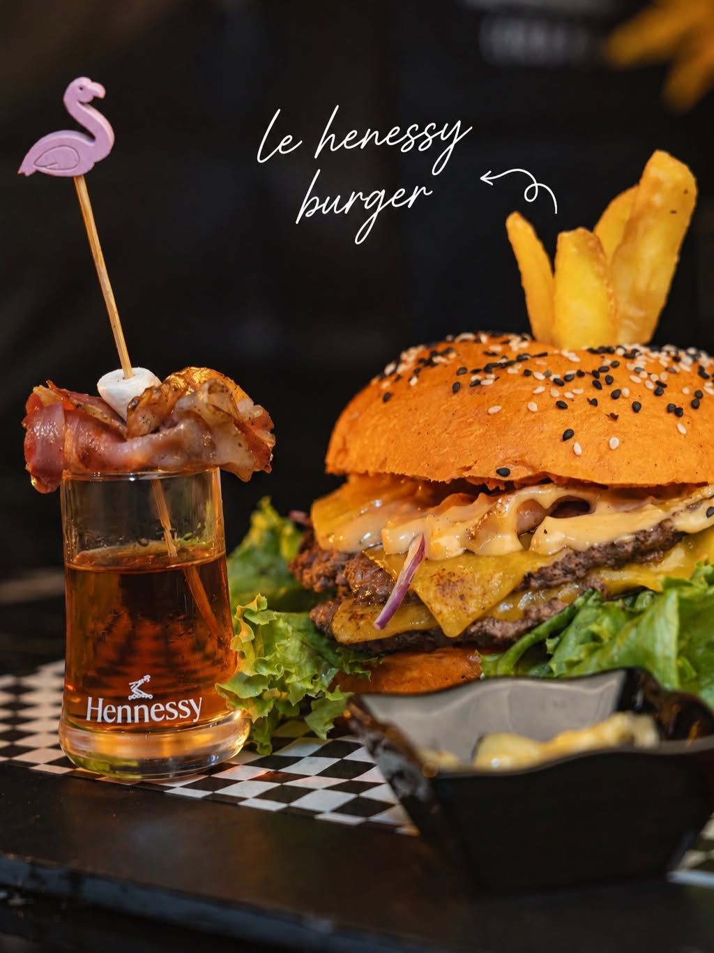 Burger et frites accompagnés d'un cocktail, servis sur une table à damier, avec l'inscription : le burger Hennessy.