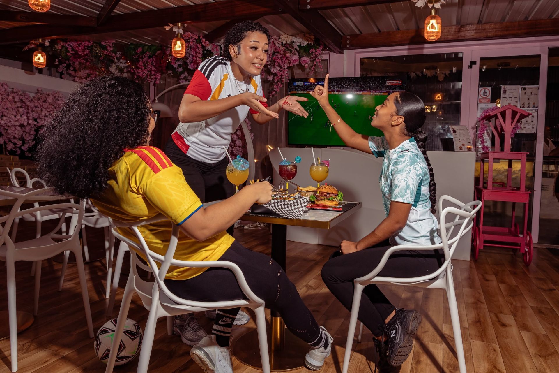 Des amis rient autour d'une table dans un restaurant à l'éclairage chaleureux, l'un d'eux se lève et fait des gestes en tendant un verre.
