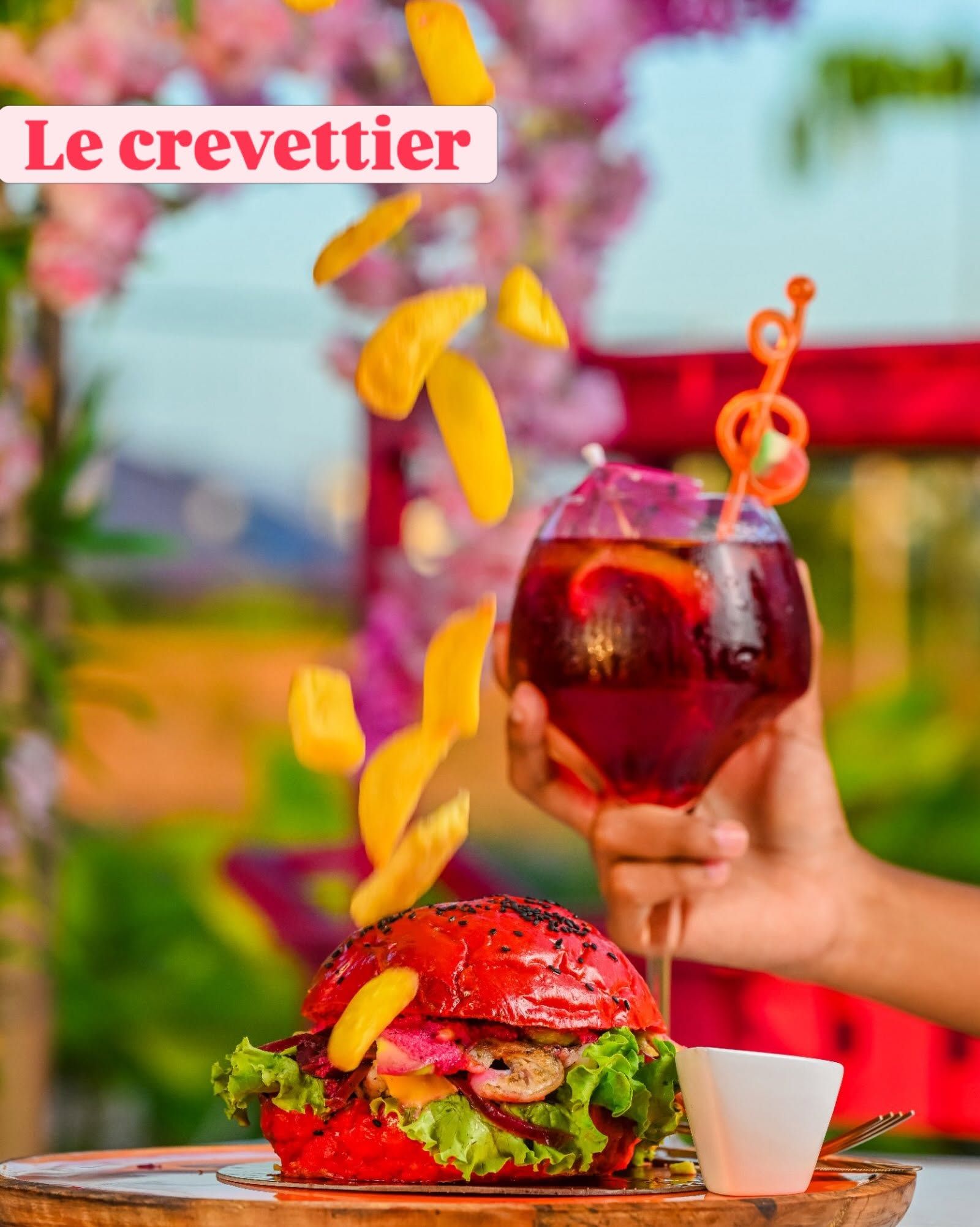 Main tenant un verre de cocktail à côté d'un hamburger gastronomique avec des frites dans un cadre extérieur coloré.