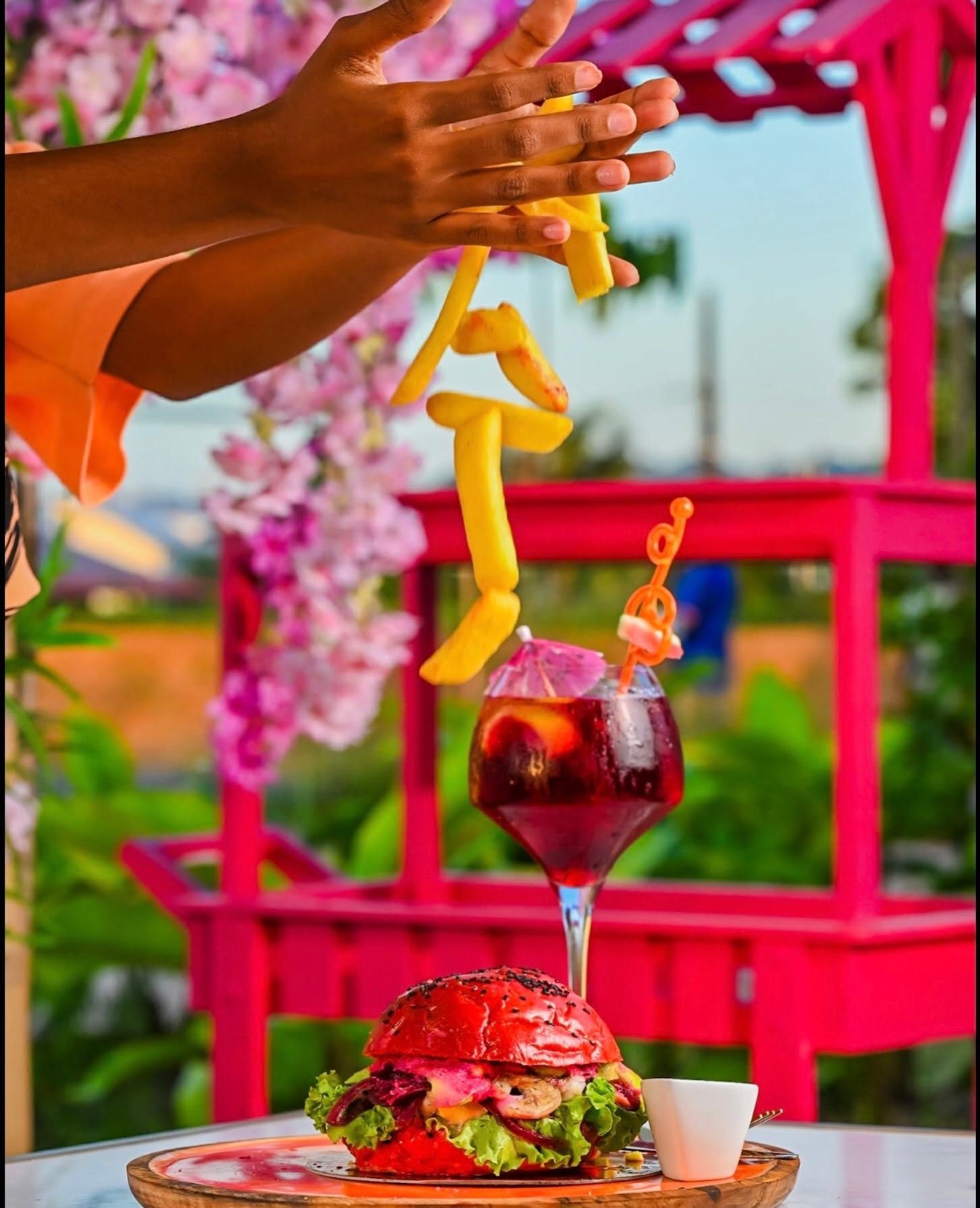 Cocktail, burger et frites sur une table rose en terrasse, avec des mains qui se tendent vers l'intérieur et un arrière-plan tropical flou.
