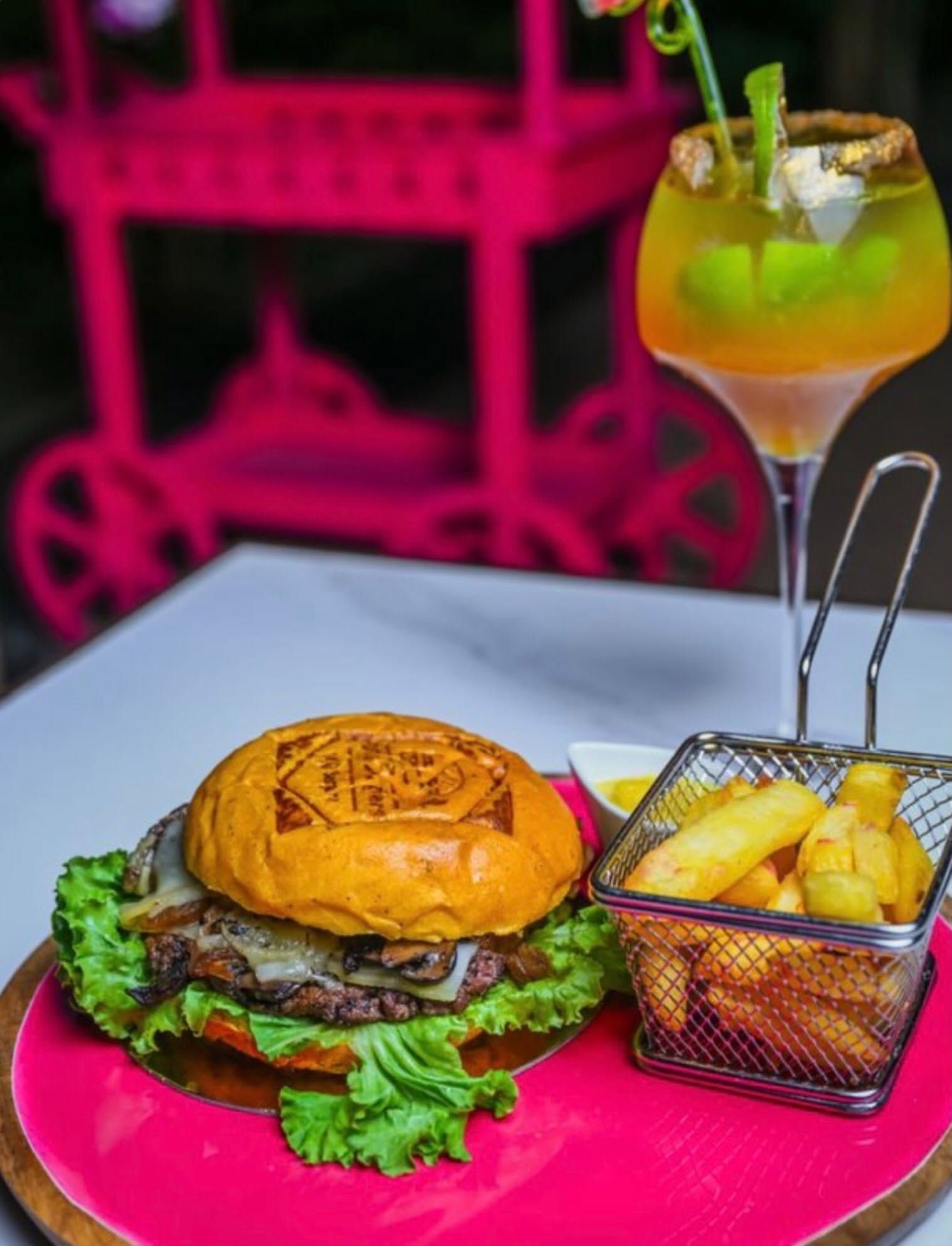 Un hamburger et des frites sur une assiette rose, à côté d'un cocktail sur une table.