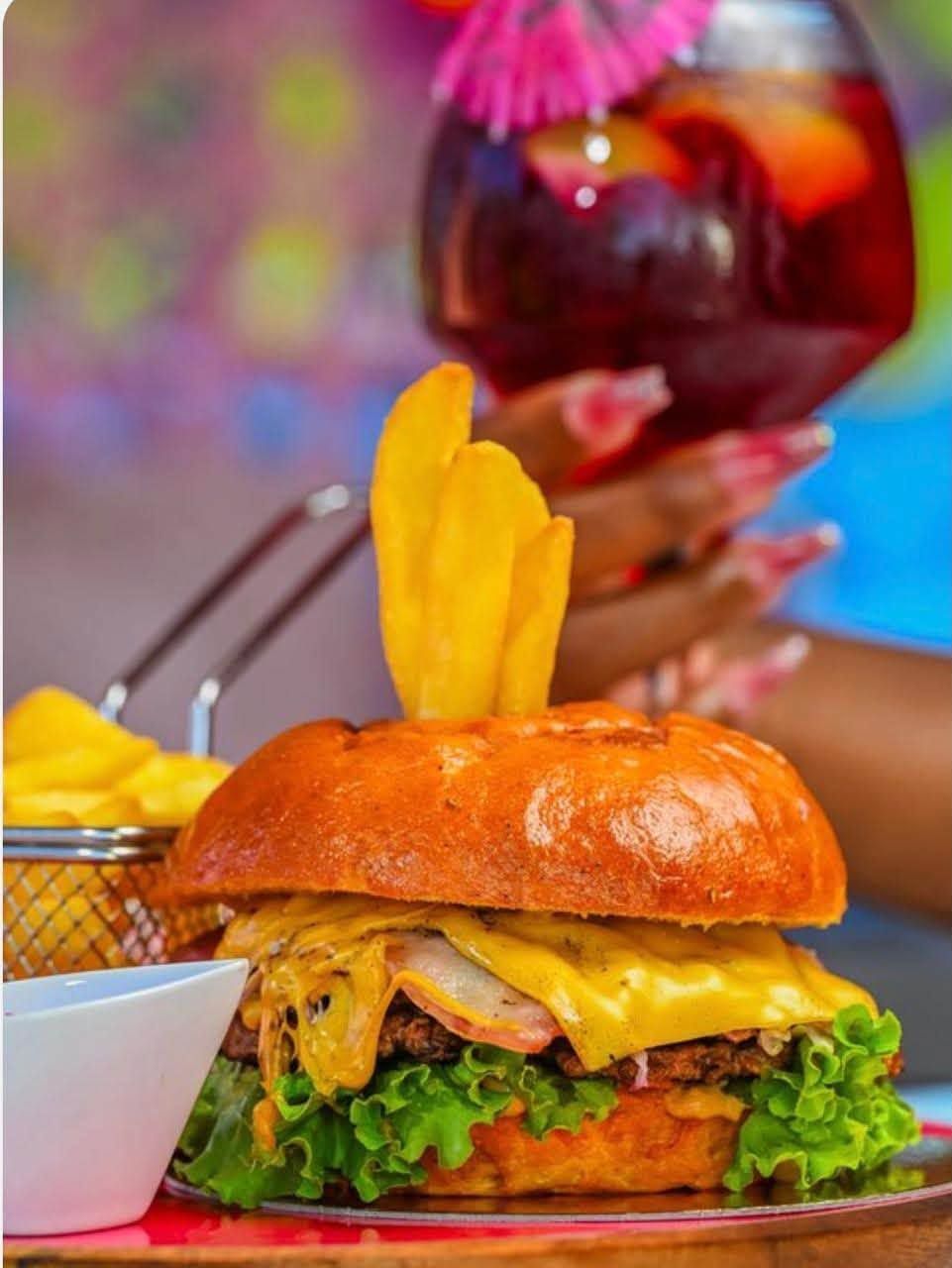 Un cheeseburger avec de la laitue et de la sauce, garni d'une garniture jaune, est posé sur une table, avec un cocktail en arrière-plan.