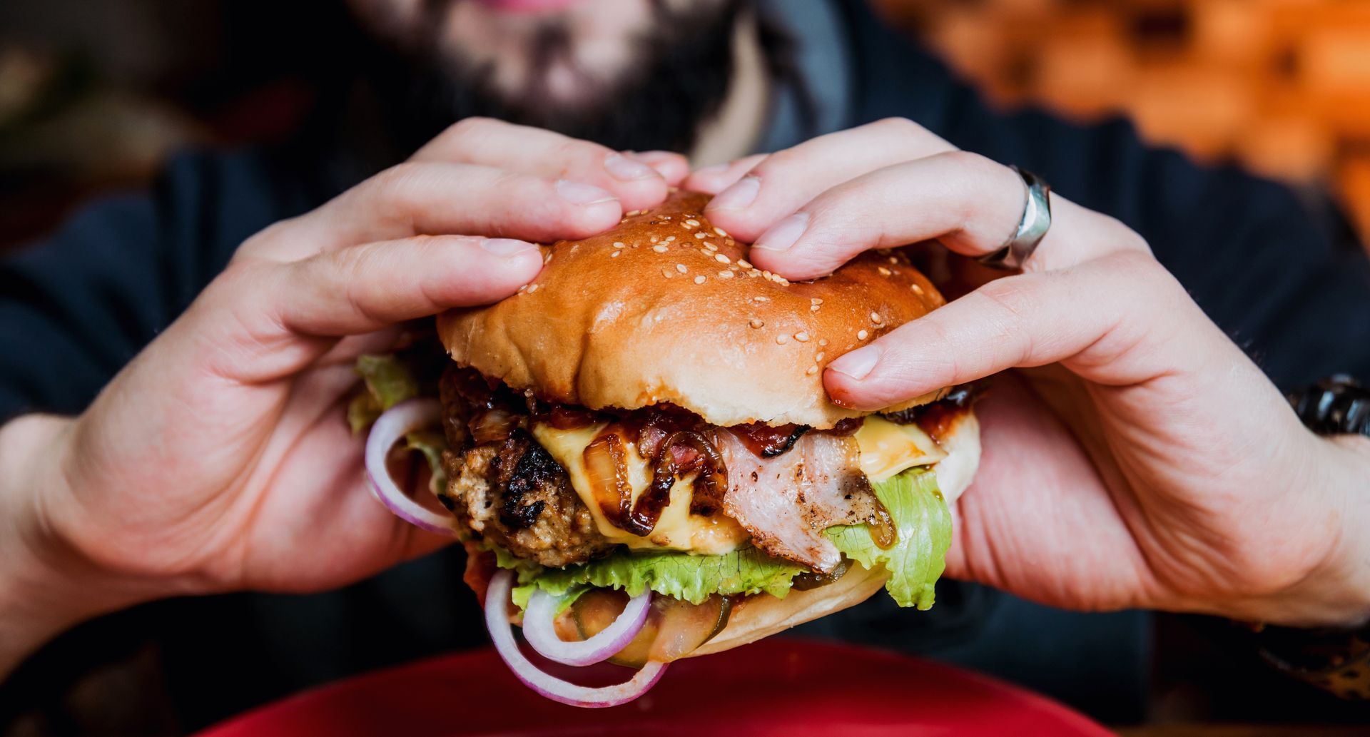 Des mains tenant un gros hamburger avec de la laitue, des oignons et du fromage fondu sur une assiette rouge.