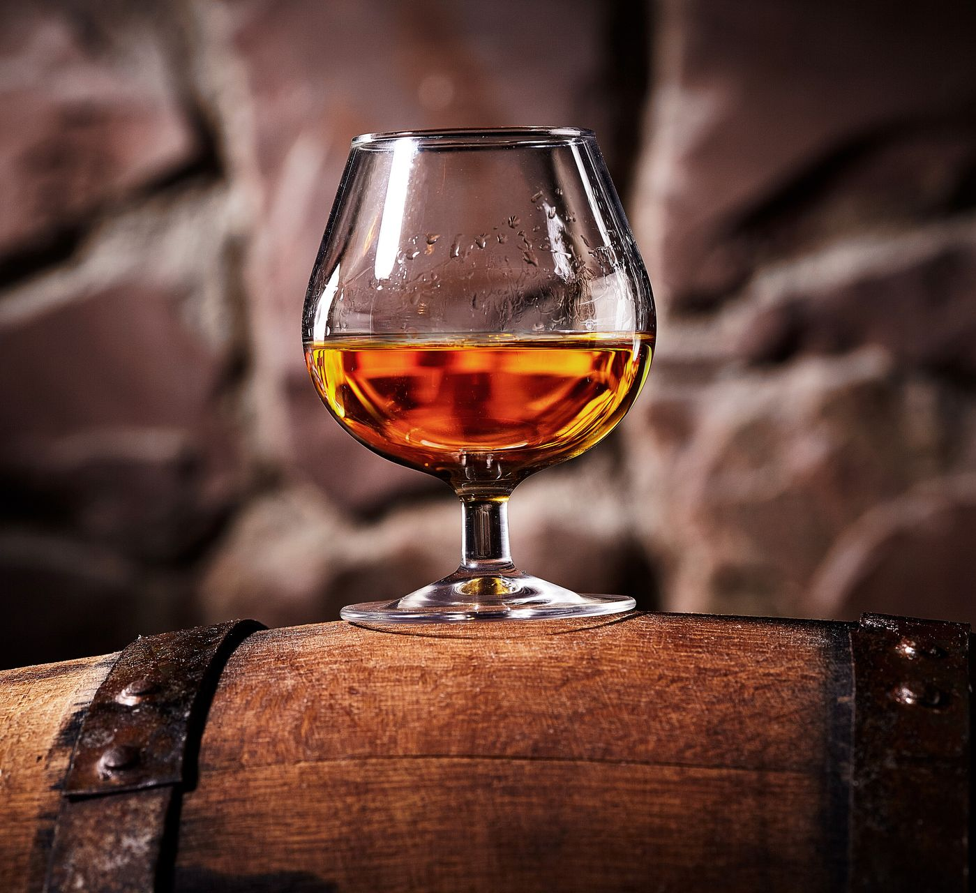 Verre de liqueur ambrée sur un tonneau en bois, devant un fond de pierre flou.