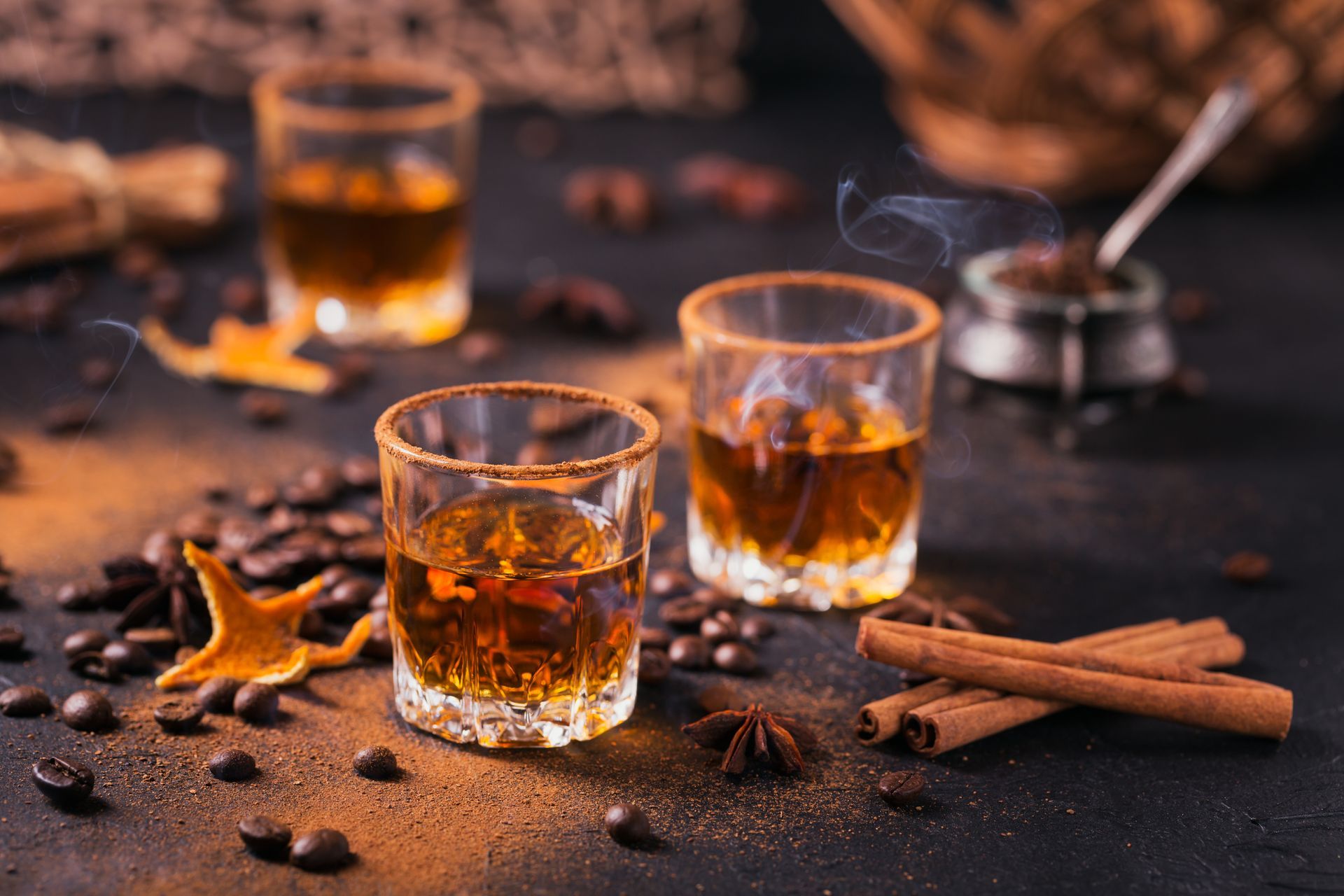 Des shots de whisky épicé sur une table sombre, avec des bâtonnets de cannelle, de l'anis étoilé et un verre chaud en arrière-plan.