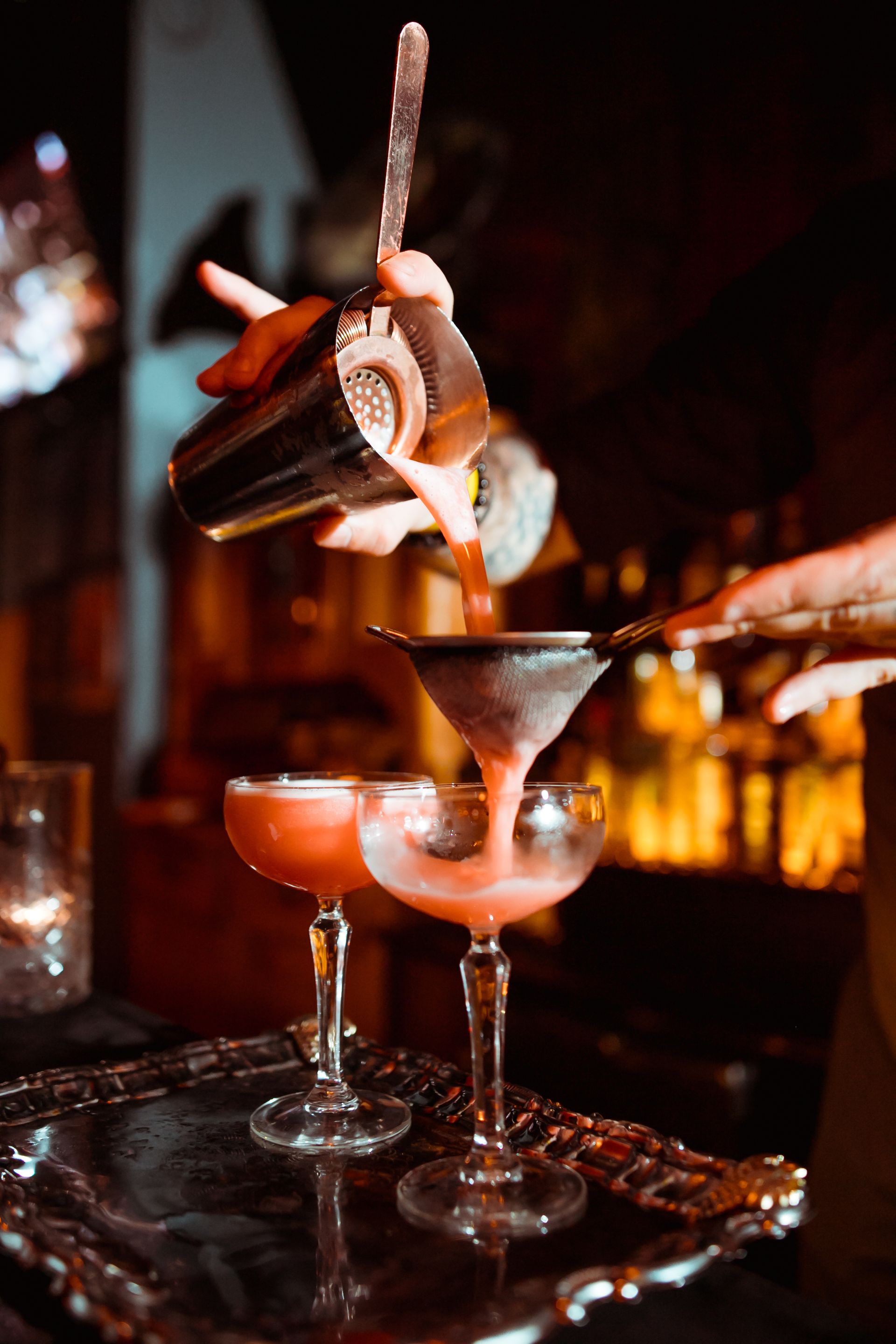 Des verres à cocktail sur un plateau, un barman prépare un verre dans un bar à l'éclairage tamisé.