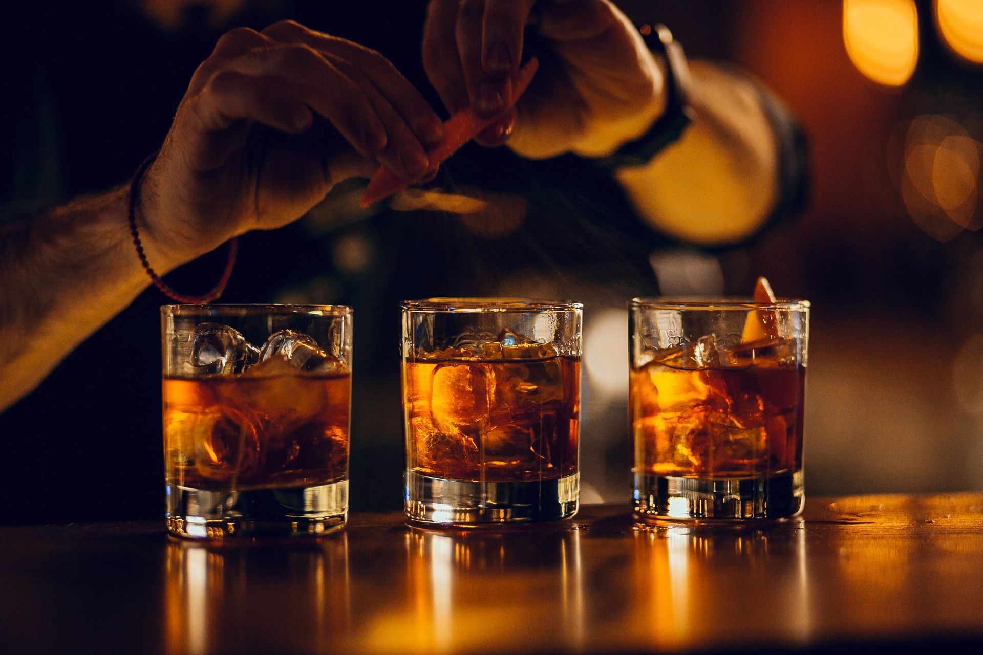 Trois verres à whisky remplis de glaçons sont posés sur un comptoir, tandis que des mains remuent les boissons sous une douce lumière ambrée.