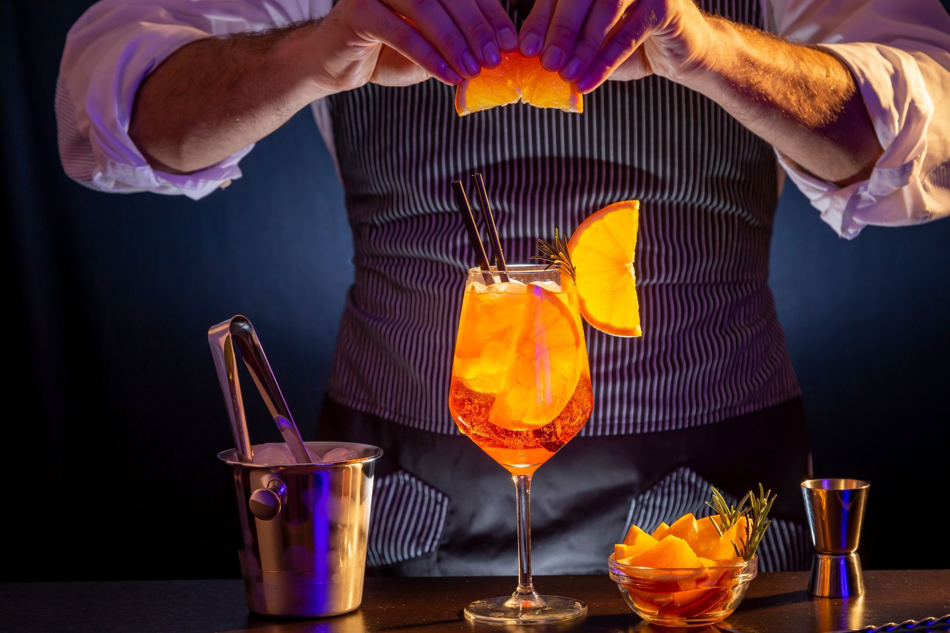 Un barman décore un cocktail orange vif avec des zestes d'agrumes violets, à côté des ustensiles de bar.