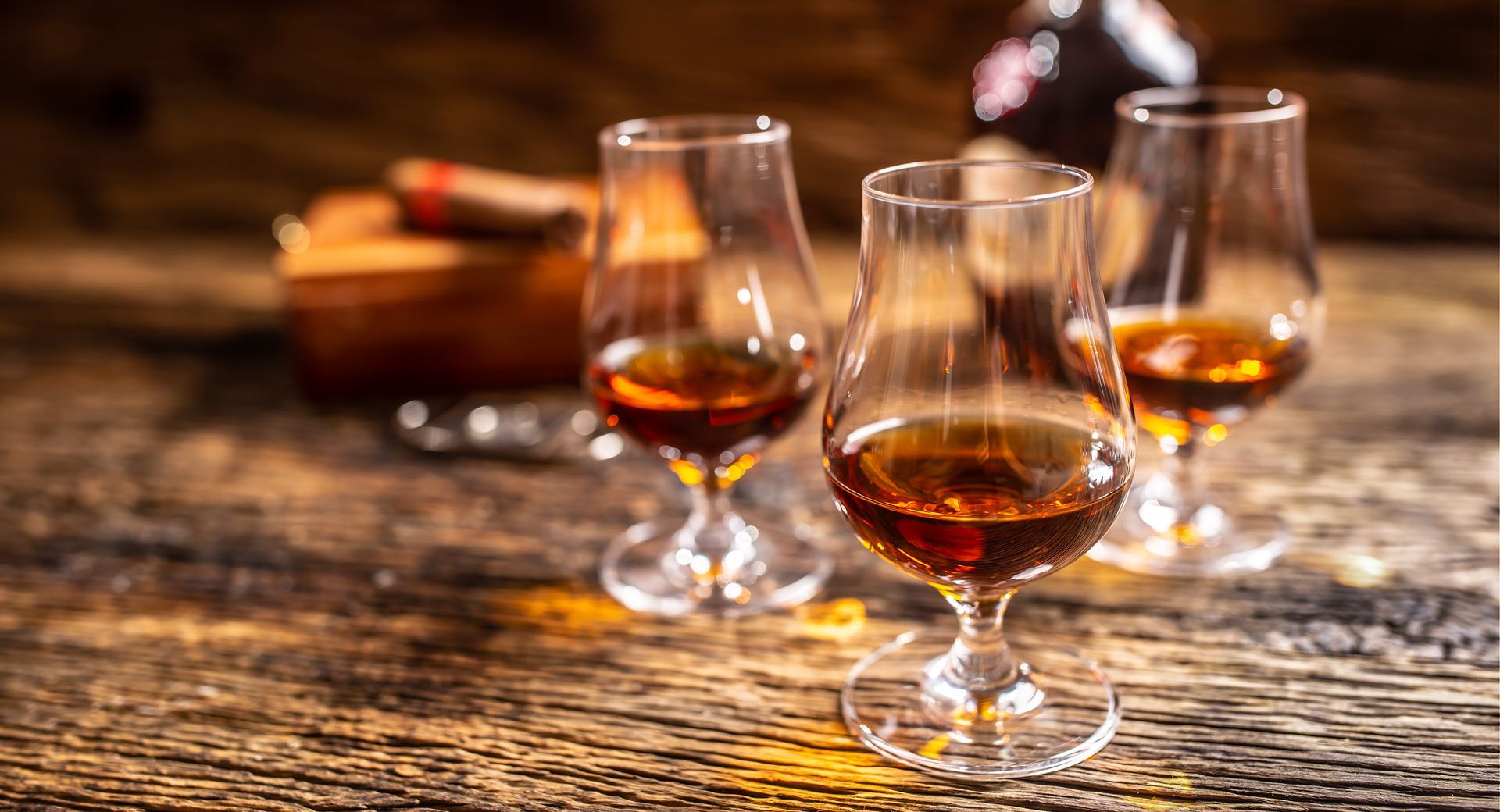 Trois verres de cognac ambré sur une table rustique en bois, avec une bouteille floue en arrière-plan.