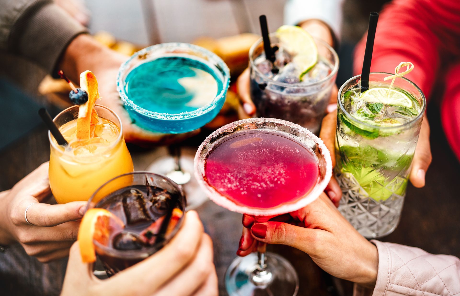 Des mains se lèvent pour trinquer avec des cocktails colorés, dont un martini bleu, un cocktail rouge et un mojito.