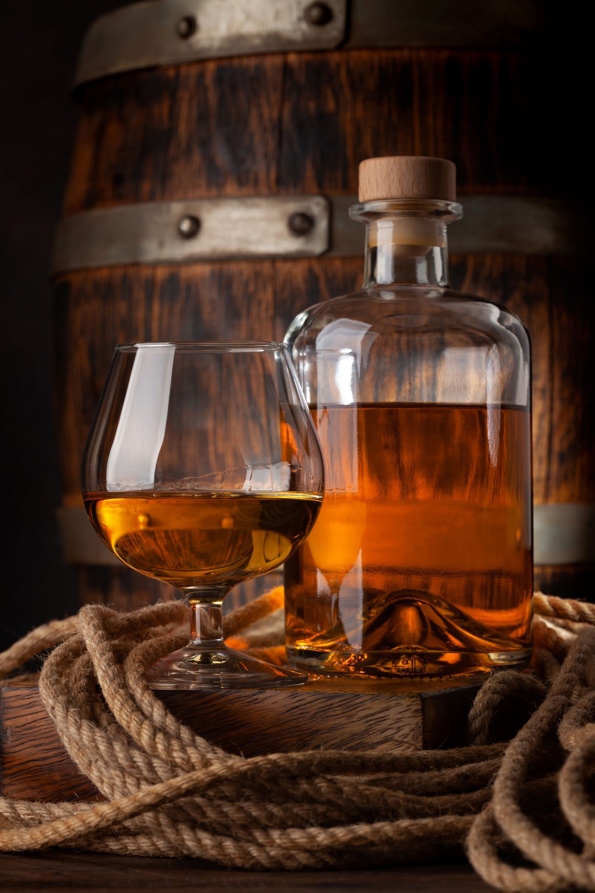 Verre et bouteille de whisky posés à une corde enroulée, devant un tonneau en bois, dans une douce lumière ambrée.