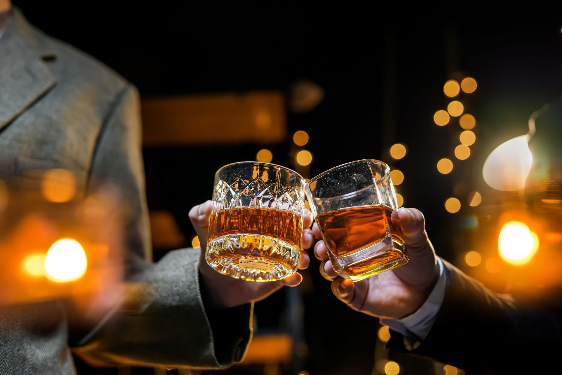 Deux mains font tinter des verres de whisky dans un bar chaleureux et tamisé, avec des lumières floues en arrière-plan.