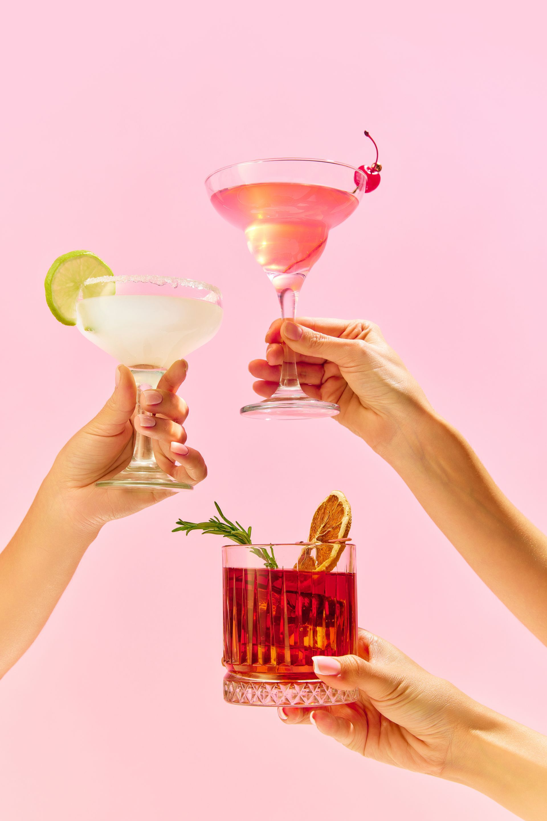 Trois mains lèvent des cocktails colorés sur un fond rose.