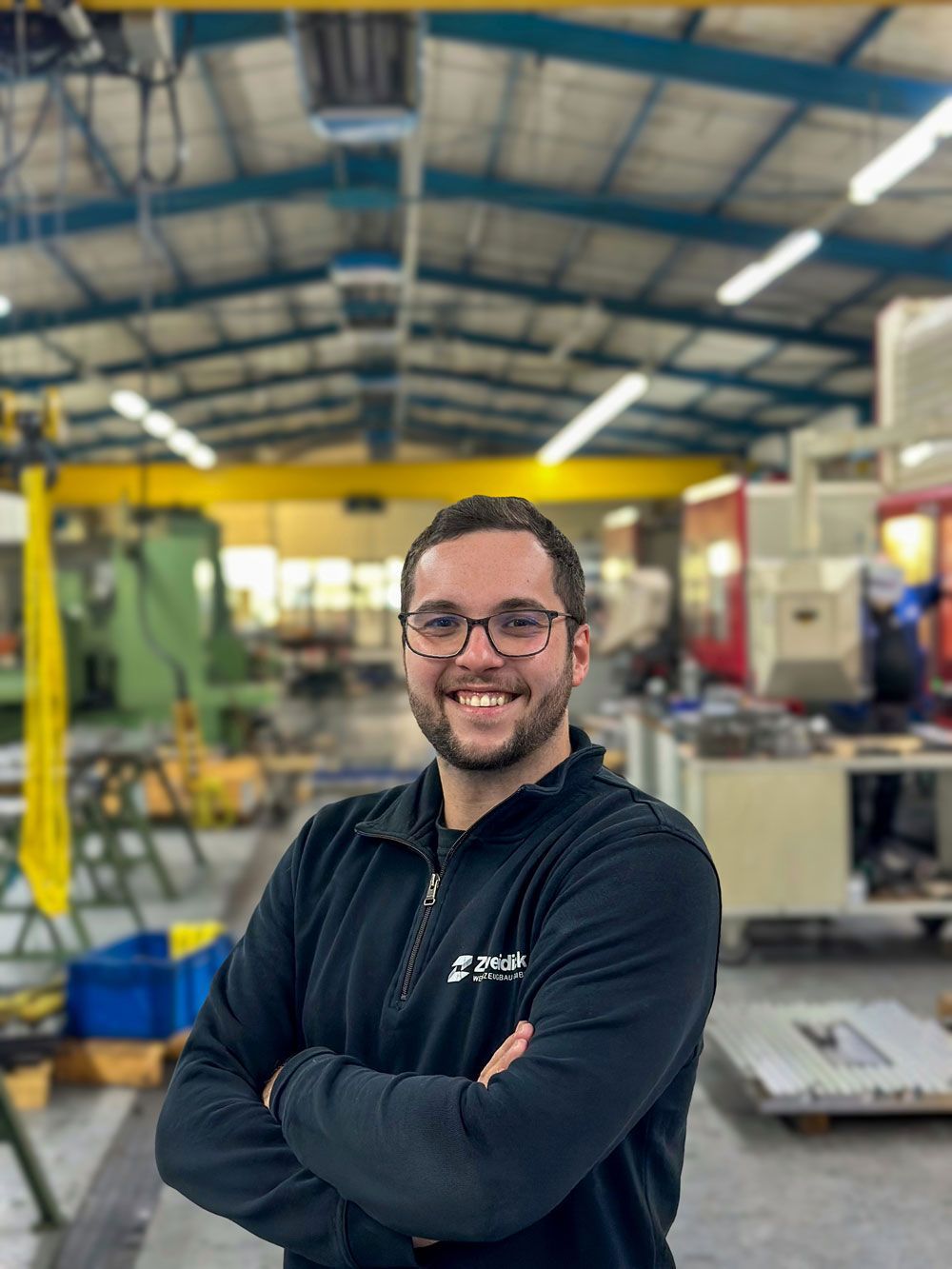 Ein lächelnder Mann mit Brille steht mit verschränkten Armen in einer Fabrik. Es ist Florian Zweidick