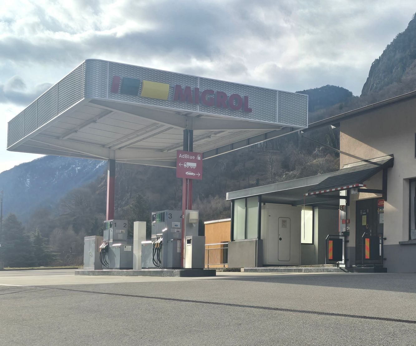 Station-service avec logo rouge et jaune sous un auvent, devant une montagne.