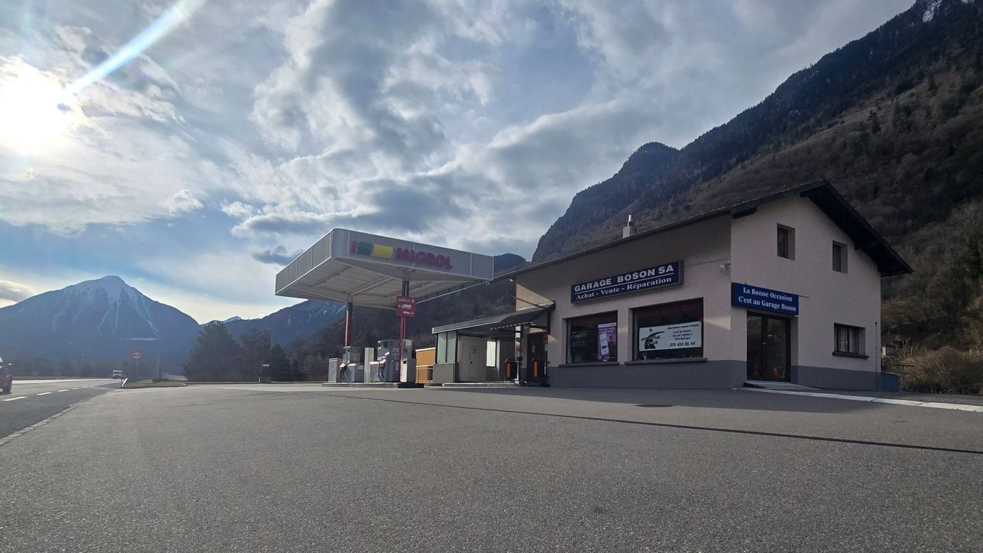 Station-service et magasin en milieu montagneux, avec une route goudronnée et un ciel nuageux.